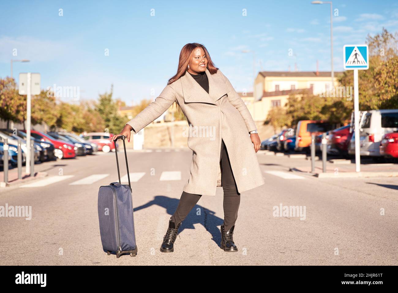 Bonne femme afro-américaine dans un pantalon noir et une veste beige tenant une valise et posant près de l'aéroport. Banque D'Images