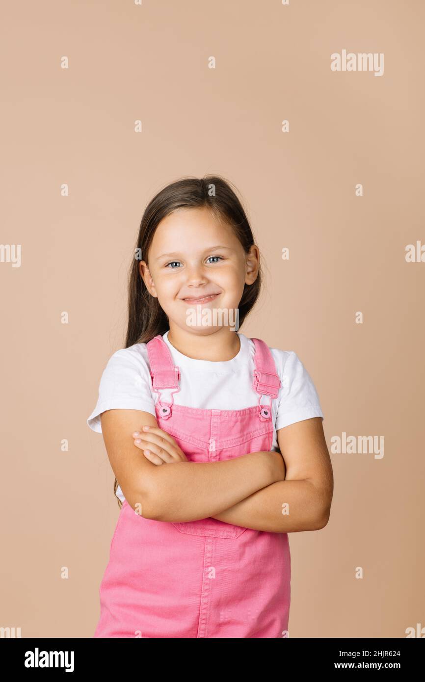 Enfant aux bras pliés avec des yeux brillants et un sourire humble regardant l'appareil photo portant une combinaison rose vif et un t-shirt blanc sur fond beige Banque D'Images