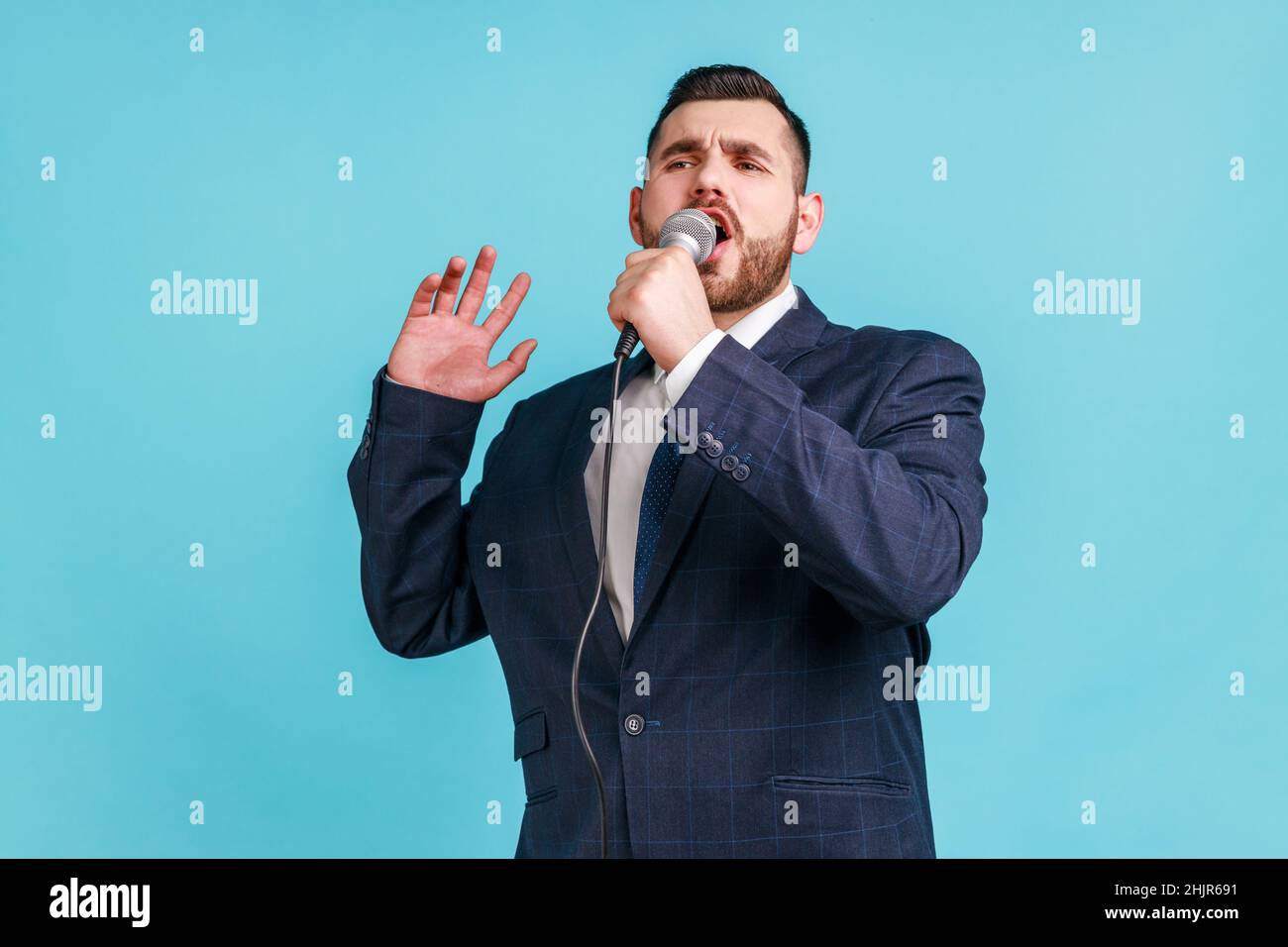 Portrait d'un jeune homme barbu adulte de talent portant un costume de style officiel chantant des chansons tenant un microphone, répétition avant la représentation.Studio d'intérieur isolé sur fond bleu. Banque D'Images