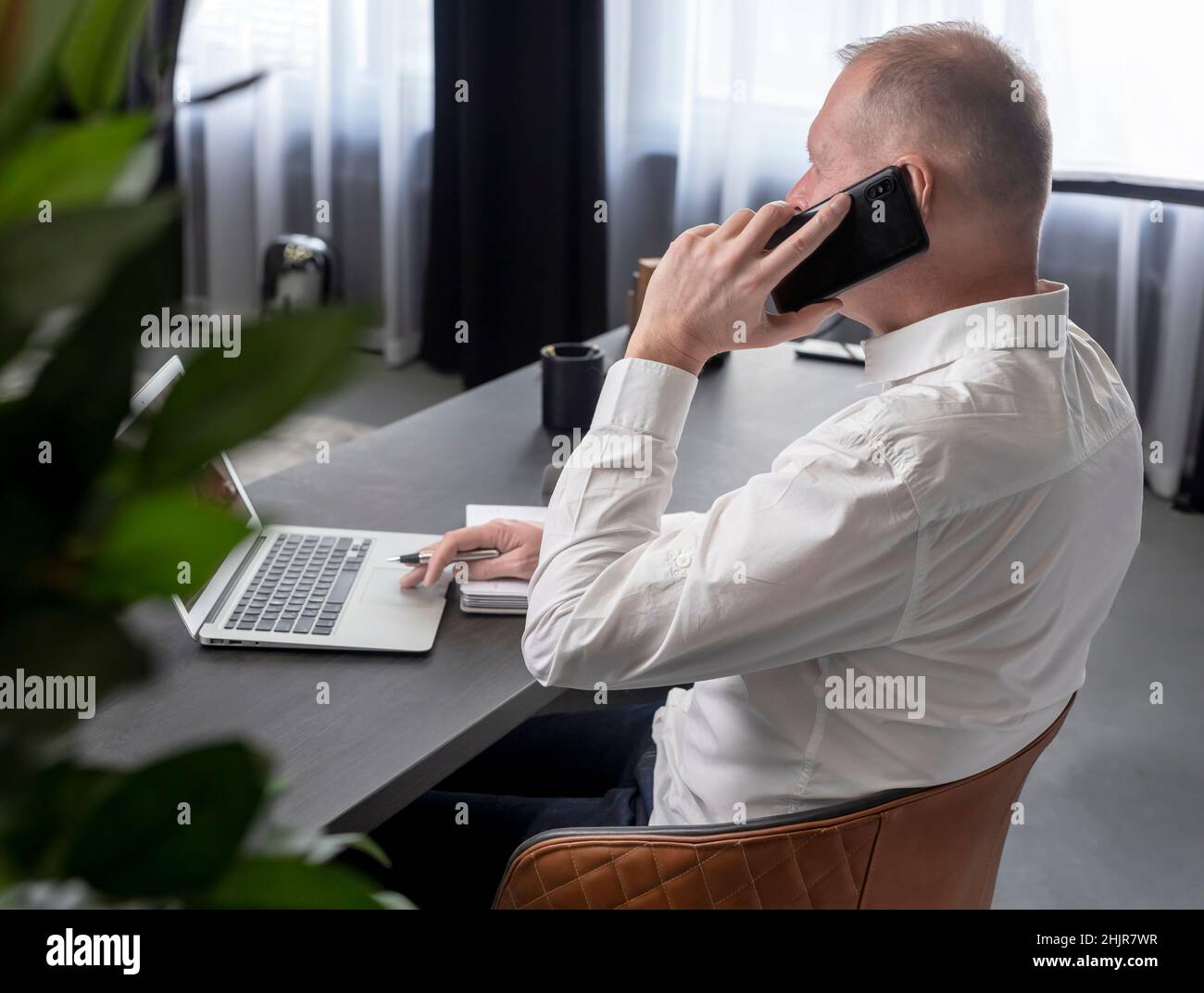 Homme d'affaires mature assis à une table au bureau, parlant sur un téléphone portable tout en regardant l'écran d'un ordinateur portable et en prenant des notes dans un ordinateur portable.Concept d'appel professionnel.Photo de haute qualité Banque D'Images