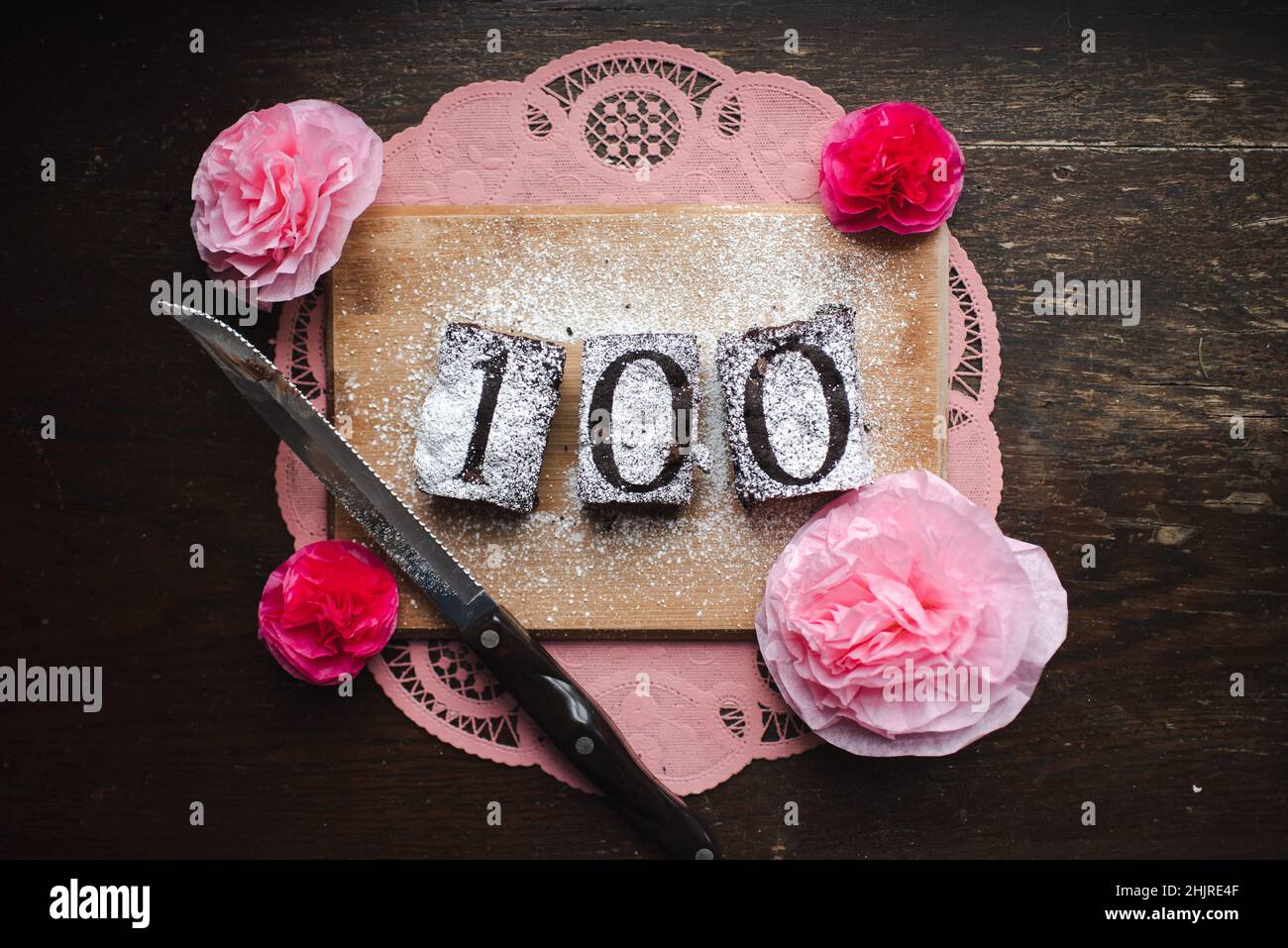 Le numéro 100 est écrit avec du sucre en poudre sur des brownies au chocolat ou un gâteau avec du napperon rose et un couteau, de la chapelure tout autour Banque D'Images