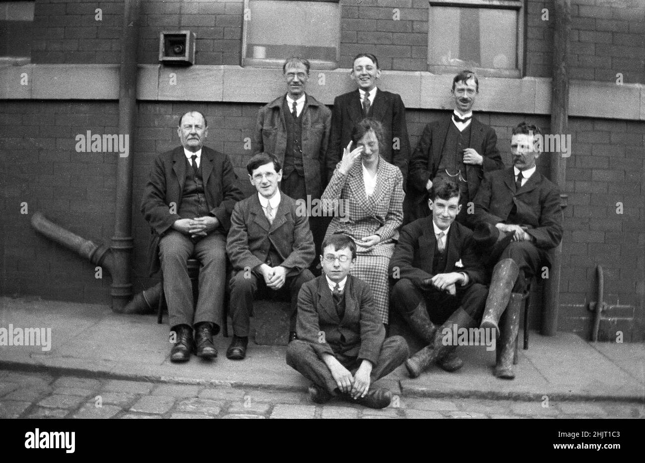 Circa 1920s, historiques, les employés de TPCG, Dudley, se rassemblent dehors sur un trottoir pour leur photo, Warwick, Angleterre, Royaume-Uni. La photo montre les vêtements de travail formels de l'époque, tous les hommes portant des costumes et des cravates. Deux des hommes ont des chaînes de montres attachées à leur gilet. La seule jeune employée est assise au milieu et s'essuie les yeux pendant qu'elle rit, avec le jeune homme derrière son échoppe riant. Le gentleman portant les bottes wellington sur la droite et avec la face arrière, pourrait être le patron, car il ne rit certainement pas! Banque D'Images