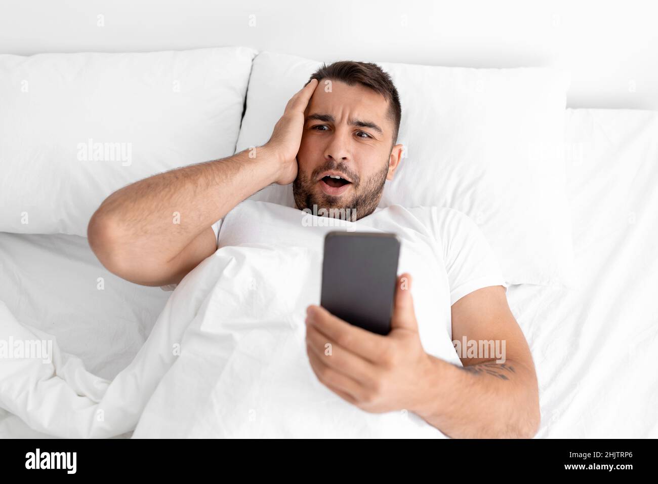 Surprise caucasien attrayant millennium gars en t-shirt blanc lit blog ou message dans le téléphone dans le lit dans la chambre Banque D'Images