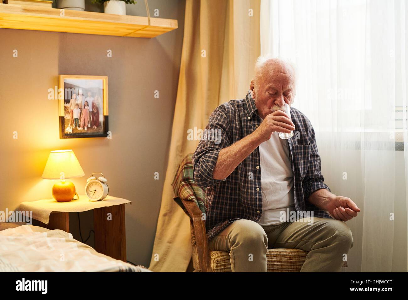 Homme senior assis sur un fauteuil dans sa chambre et de l'eau potable, du verre, il prend des pilules le matin Banque D'Images