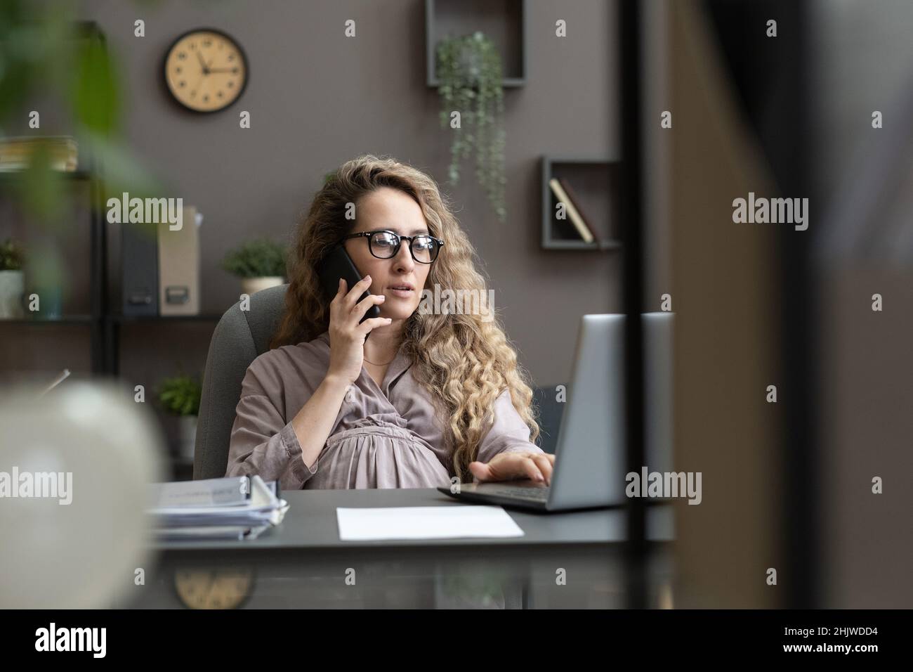 Jeune femme enceinte en robe grise décontractée parlant au téléphone portable tout en étant assise devant un ordinateur portable et en regardant l'affichage Banque D'Images