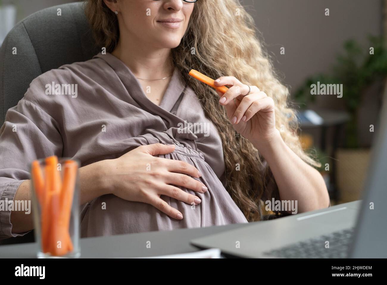 Femme d'affaires enceinte contemporaine mangeant des carottes et gardant la main sur le ventre tout en étant assise devant un ordinateur portable Banque D'Images