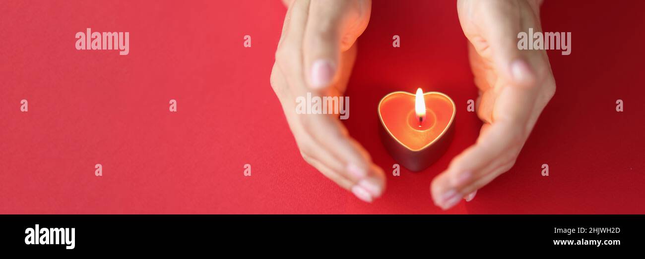 La main femelle protège soigneusement la bougie brûlante en forme de coeur sur fond rouge Banque D'Images