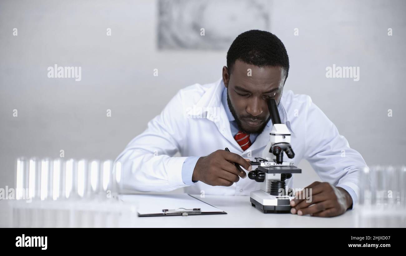 scientifique afro-américain regardant à travers le microscope près du presse-papiers en laboratoire Banque D'Images