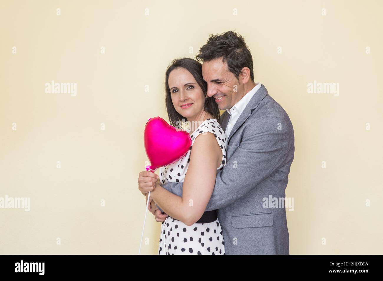 Vue latérale d'un homme hispanique souriant embrassant une femme élégante avec un ballon en forme de cœur depuis l'arrière tout en montrant le concept de la Saint-Valentin sur un baba beige Banque D'Images