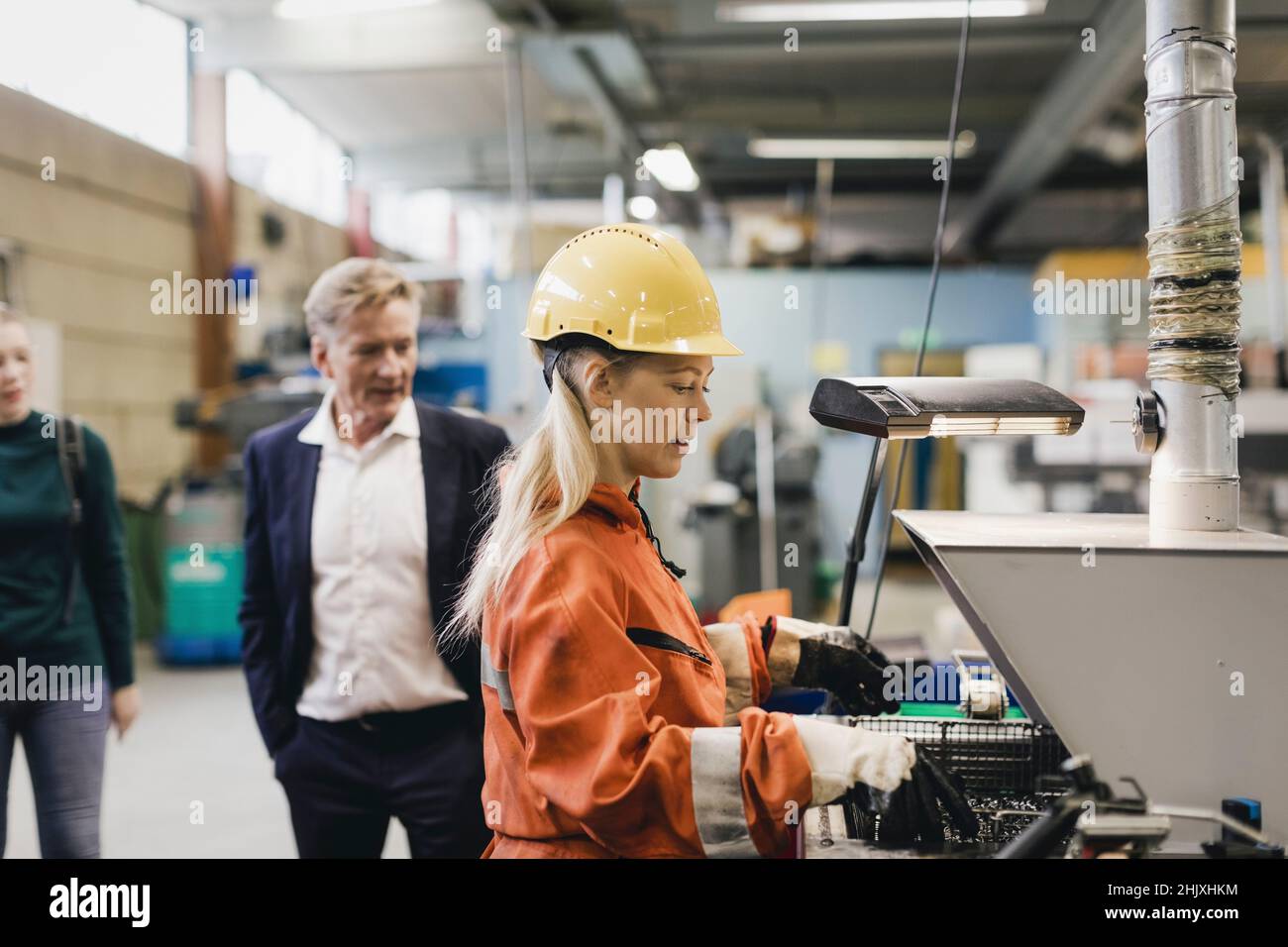 Employée industrielle travaillant dans hardhat sur des machines pendant que les directeurs en arrière-plan à l'usine Banque D'Images
