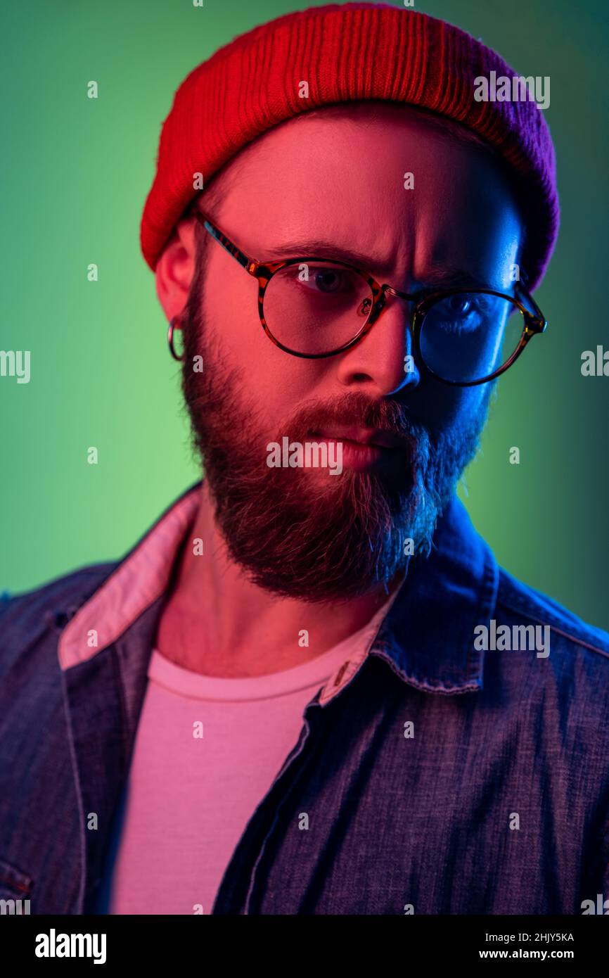 Portrait en gros plan de l'homme de taille basse pensif en lunettes, se tenant à l'écart, exprimant sa prévenance, portant un bonnet beanie et une veste en denim.Studio d'intérieur isolé sur fond de néon coloré. Banque D'Images
