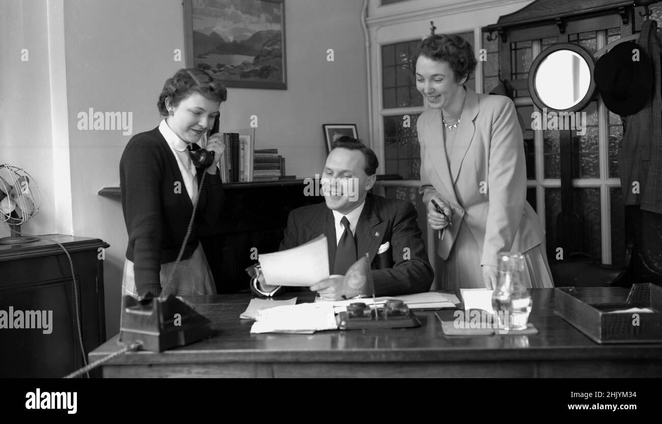 1950s, historique, homme d'affaires adapté à son bureau, avec sa secrétaire féminine au téléphone et une assistante personnelle féminine élégamment habillée, debout à côté de lui, avec un stylo plume à la main pour signer des lettres, Angleterre, Royaume-Uni. Banque D'Images