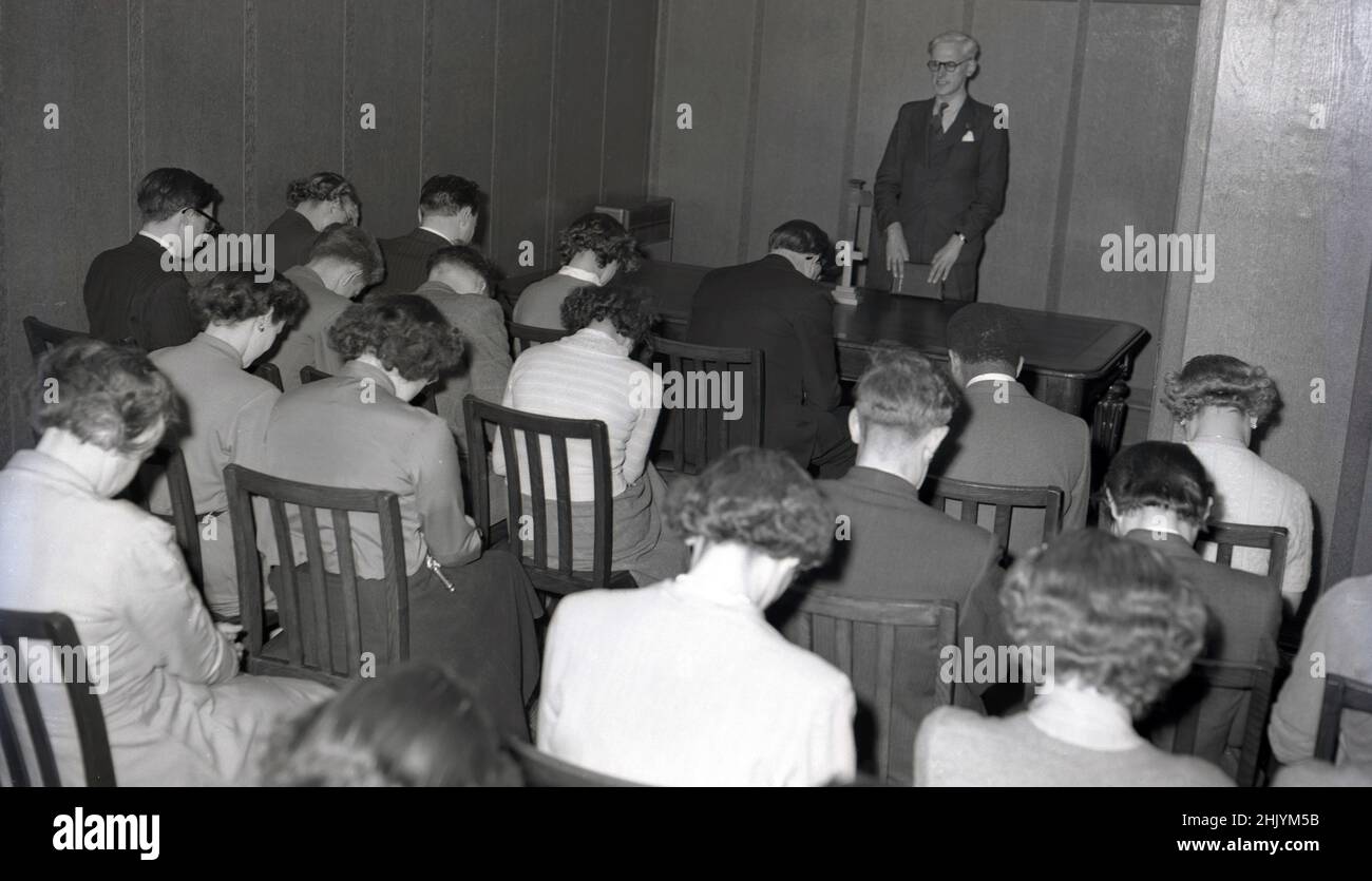 1950s, historique, un service chrétien.. un certain nombre d'hommes et de femmes assis sur des chaises en bois à l'intérieur d'une petite pièce, les têtes badient, dans la prière.Un pasteur masculin dans un costume et une cravate et tenant une bible se tient derrière une table avec une petite croix sur face à sa congrégation, USA. Banque D'Images