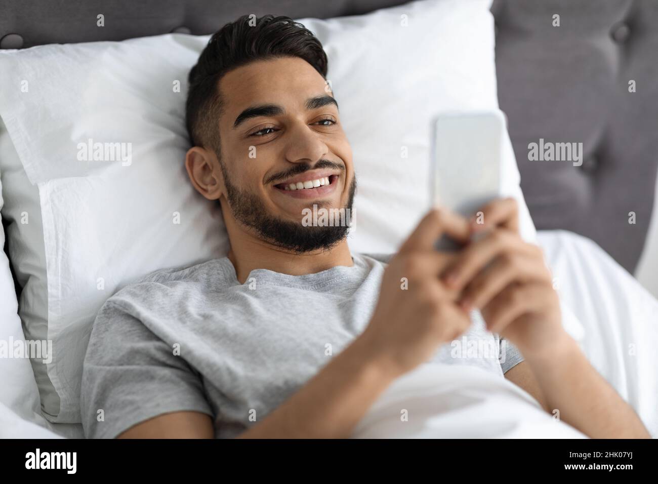 Jeux mobiles.Joyeux jeune homme arabe utilisant un smartphone tout en étant allongé dans le lit Banque D'Images