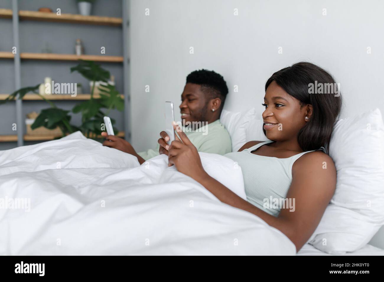 Rire jeune homme noir et femme bavardant sur les smartphones se trouvent sur le lit blanc dans la chambre Banque D'Images