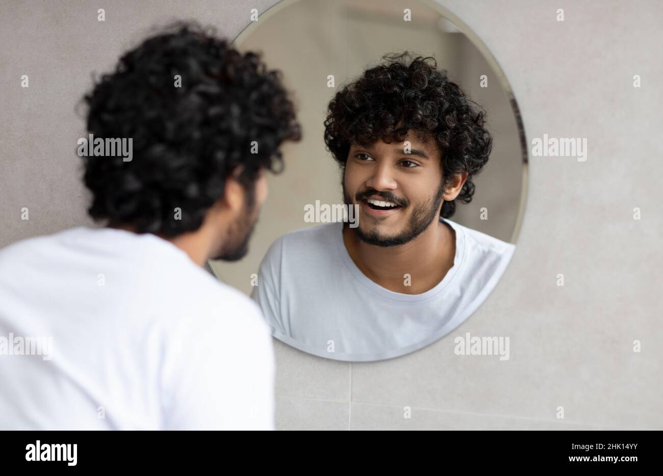 Portrait d'un Indien heureux regardant son reflet dans le miroir rond sur le mur, jeune homme debout dans la salle de bains Banque D'Images