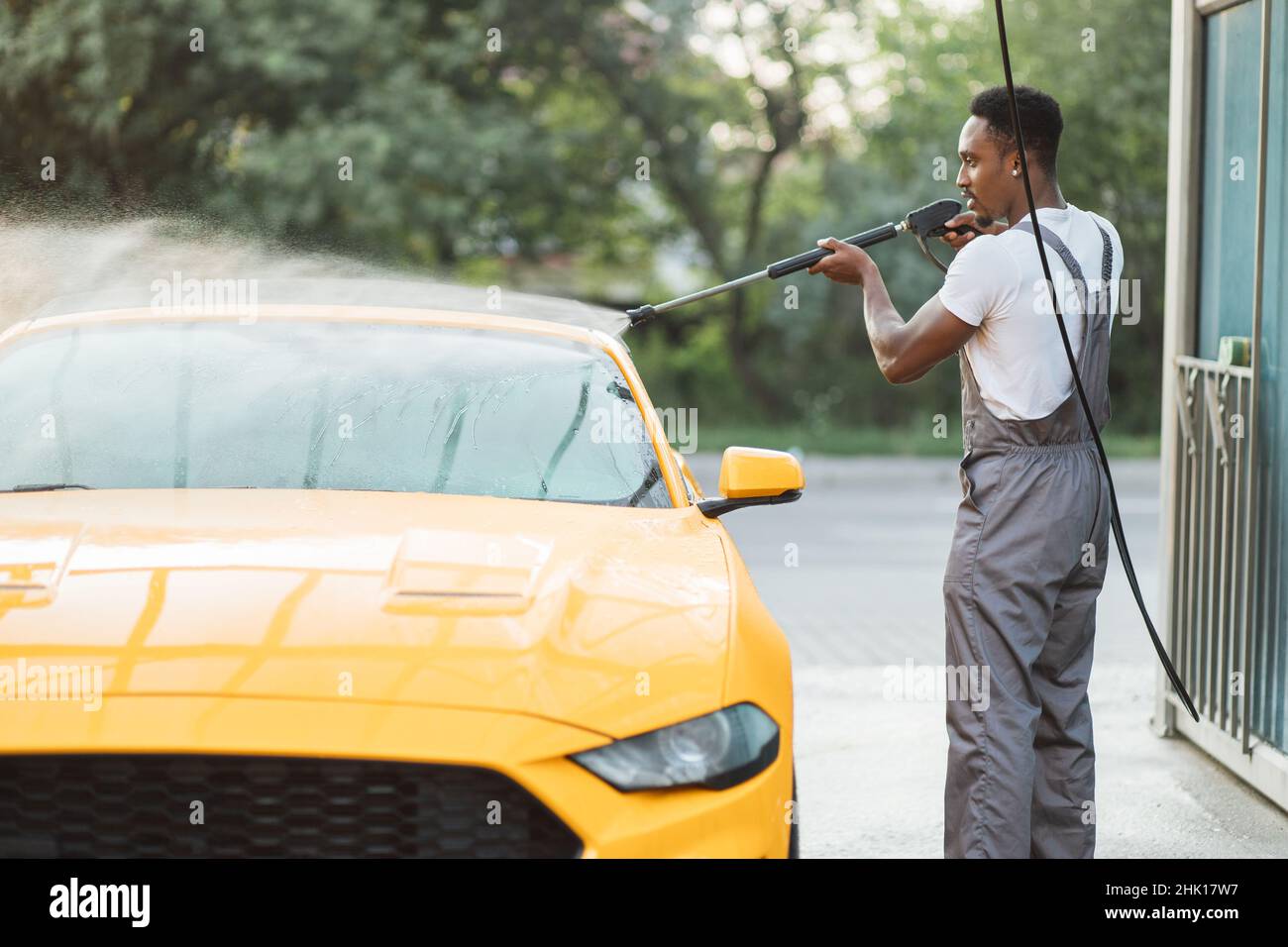 Lavage manuel de la voiture.Beau jeune homme africain lavant son véhicule jaune de luxe avec pompe à eau haute pression à l'automobile de nettoyage libre-service à l'extérieur. Banque D'Images