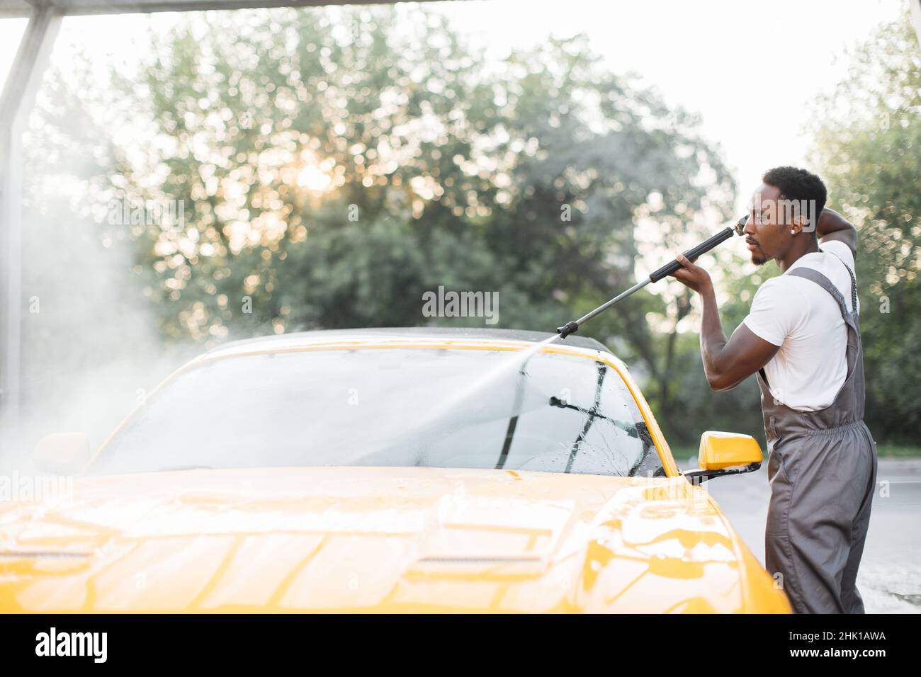 Lavage manuel de la voiture.Beau jeune homme africain lavant son véhicule jaune de luxe avec pompe à eau haute pression à l'automobile de nettoyage libre-service à l'extérieur. Banque D'Images