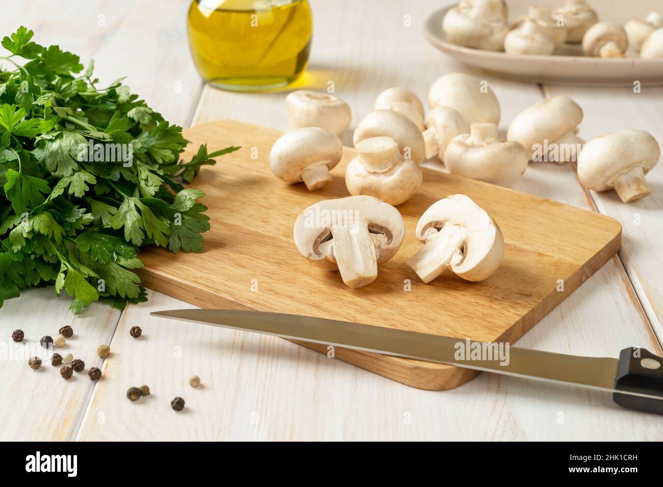 Champignon cru de moitié et couteau de cuisine sur une planche à découper, bouquet de persil et champignons de Paris entiers sur une table en bois blanc.Cuisson des champignons. Banque D'Images