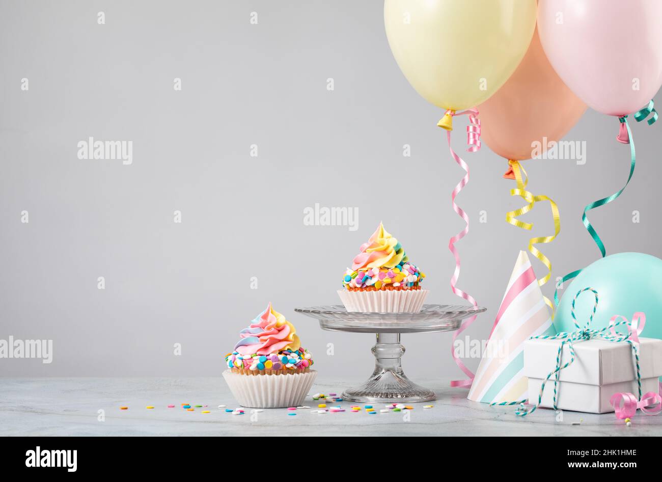 Deux petits gâteaux d'anniversaire arc-en-ciel avec cadeaux, chapeaux et ballons colorés sur fond gris clair.Scène d'une fête d'anniversaire! Banque D'Images