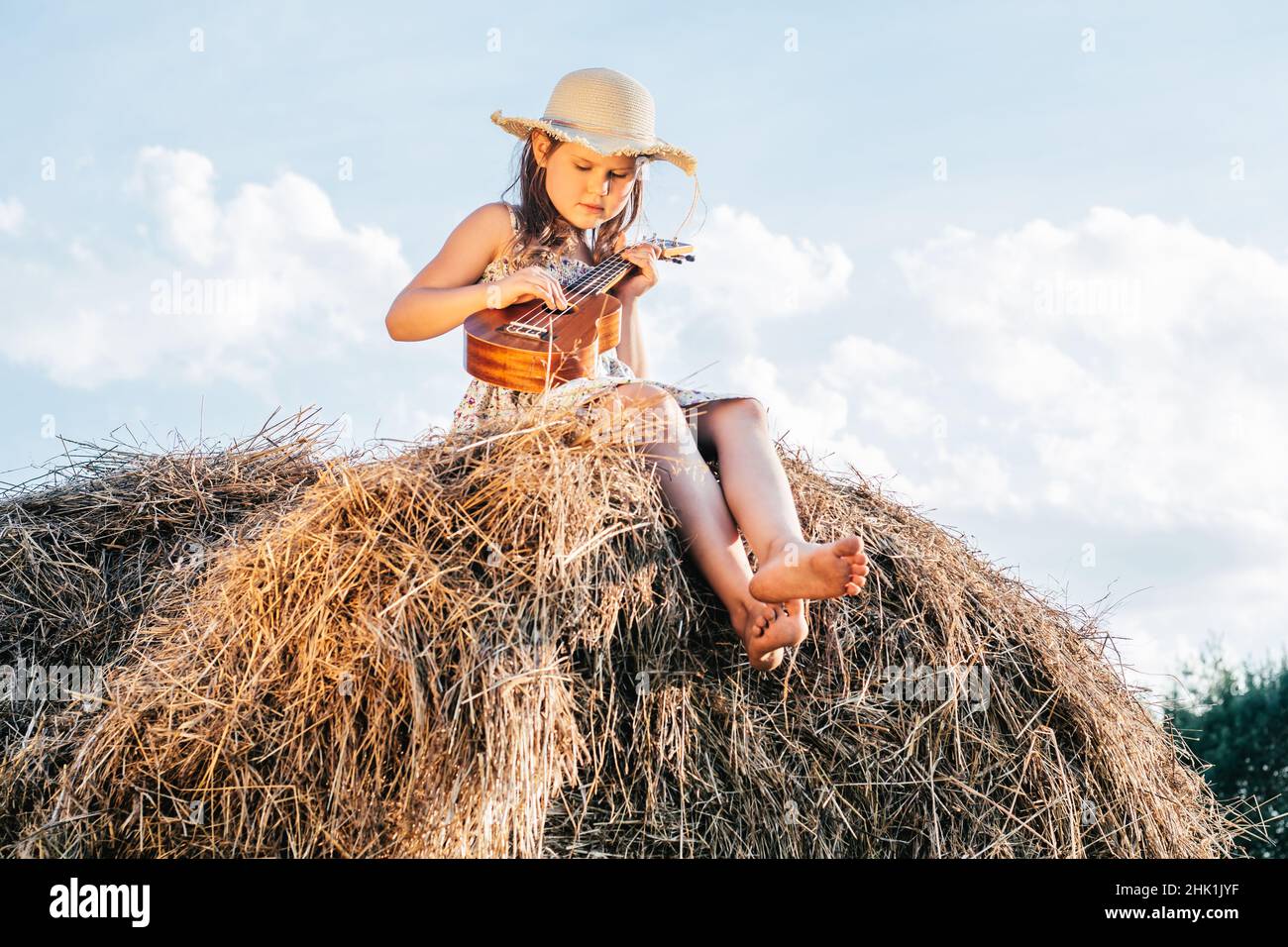 Portrait de petite fille jouant de la petite guitare ukulele, plongeant sur haystack dans le champ.Jour nuageux et ensoleillé.Concept joyeux et agréable.Arbres à l'arrière Banque D'Images