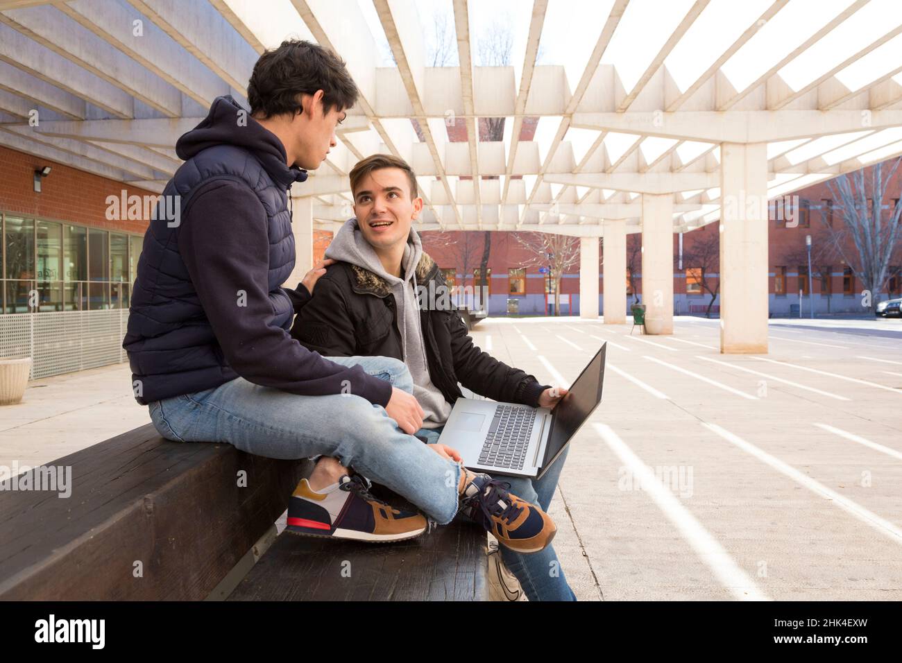 vue latérale deux amis étudiants assis sur un banc avec un ordinateur portable regardant l'un l'autre Banque D'Images