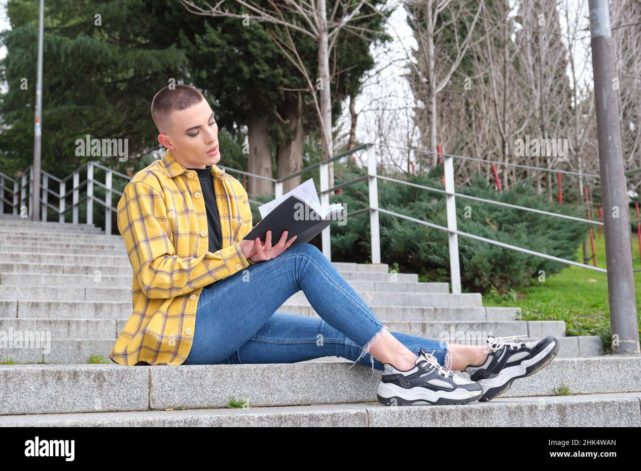 Jeune personne non binaire assis sur les escaliers et lisant un livre. Banque D'Images