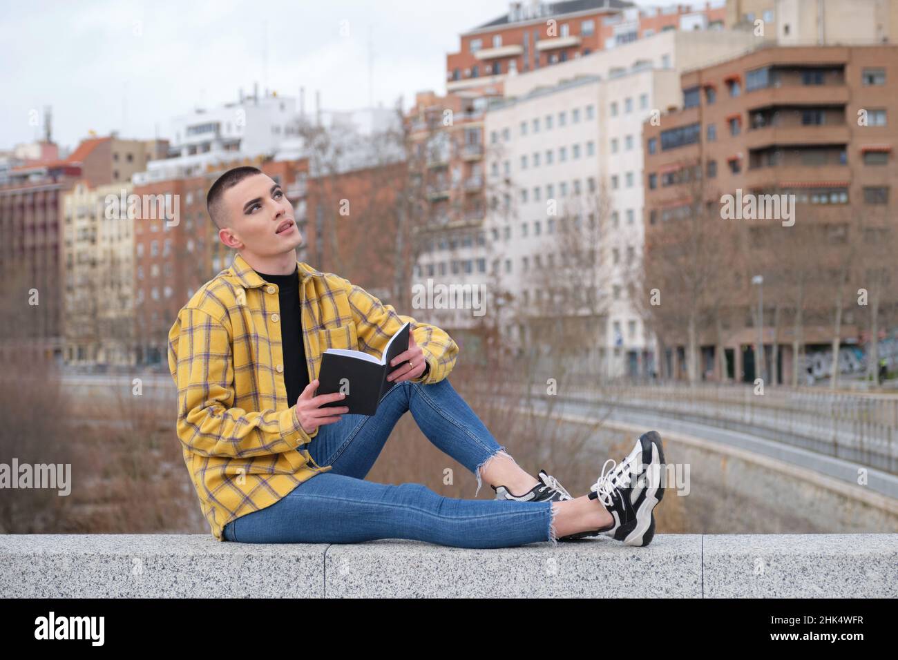 Jeune personne non binaire assis sur un mur et lisant un livre. Banque D'Images