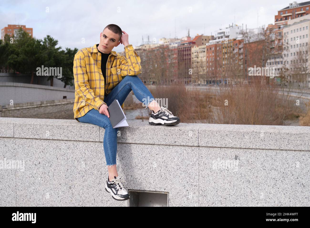 Jeune personne non binaire assis sur un mur avec un livre regardant la caméra. Banque D'Images