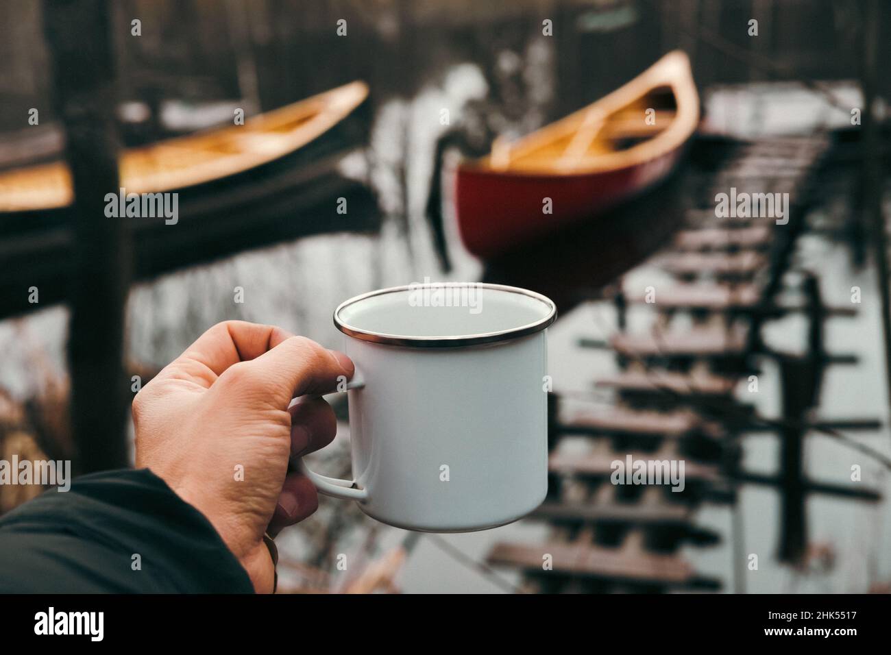Une tasse de randonnée en étain à la main avec des bateaux de canoë en arrière-plan, point de vue.Pagayer au printemps ou en automne Banque D'Images