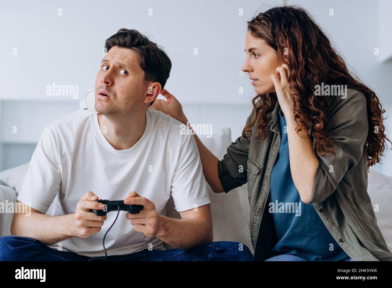 Une femme inquiète vient à son mari dans un casque sans fil blanc jouant à des jeux informatiques avec enthousiasme et querelles et se bat pour arrêter le joueur Banque D'Images