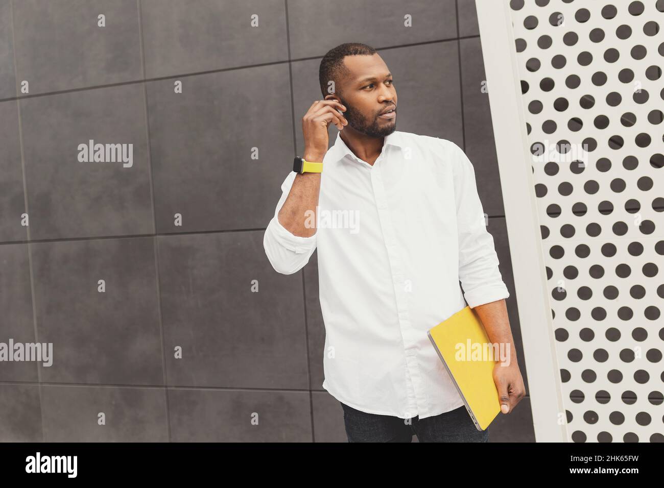 Portrait d'un indépendant habillé et élégant, debout près du bureau, mettant des écouteurs pour écouter de la musique. Homme d'affaires afro-américain tenant un ordinateur portable Banque D'Images