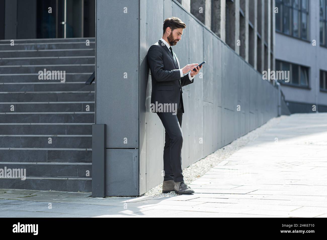 Employé caucasien barbu sur fond urbain immeuble de bureau texting messages, lire les bonnes nouvelles téléphone mobile.Homme d'affaires debout dans un désordre de costume formel Banque D'Images