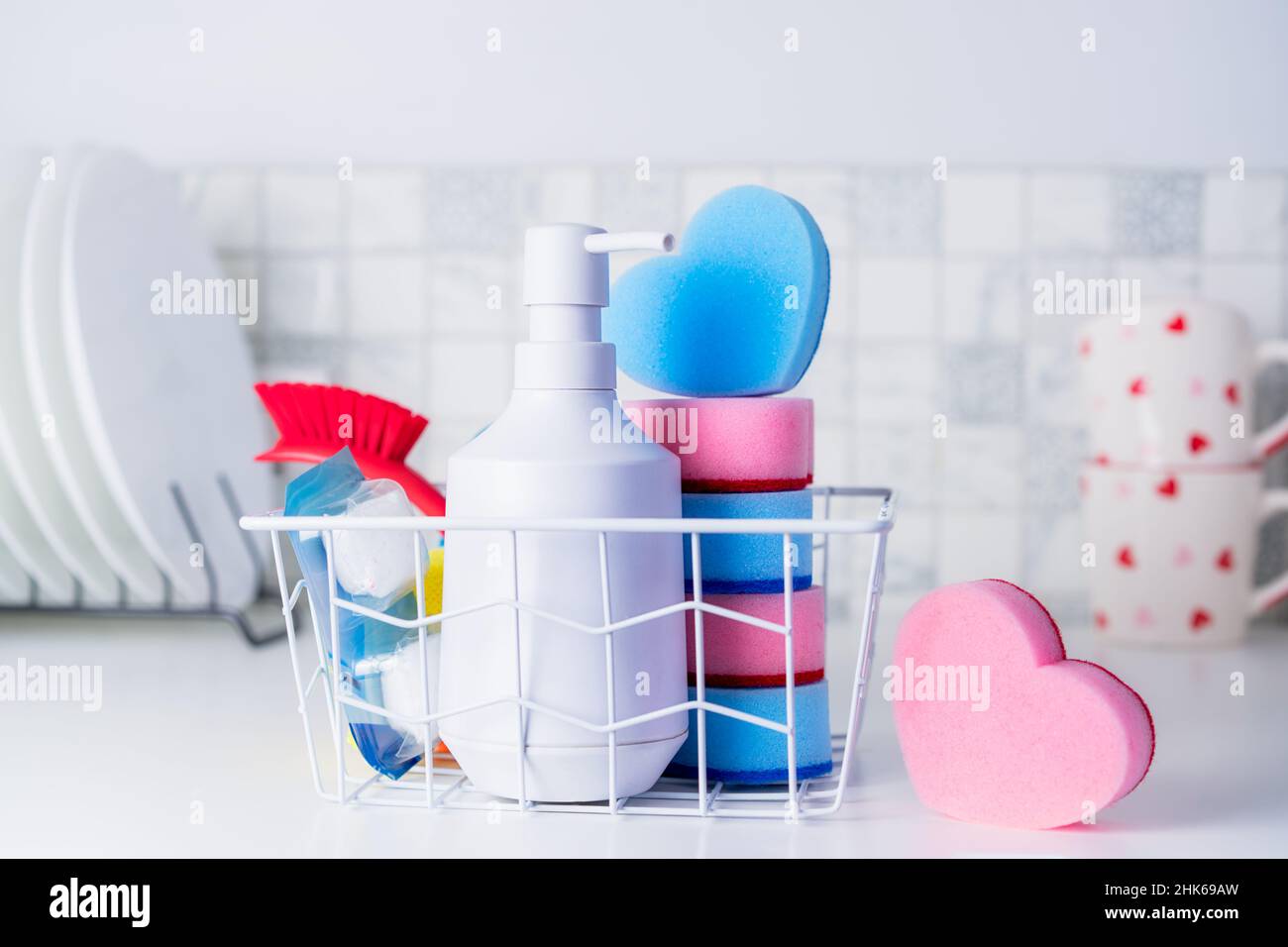 Concept d'entretien ménager.Savon à vaisselle, éponges en forme de coeur, brosse et pilules dans le panier sur le fond des assiettes et tasses propres sur la table de cuisine. Banque D'Images Concept d'entretien ménager.Savon à vaisselle, éponges en forme de coeur, brosse et pilules dans le panier sur le fond des assiettes et tasses propres sur la table de cuisine. Banque D'Images