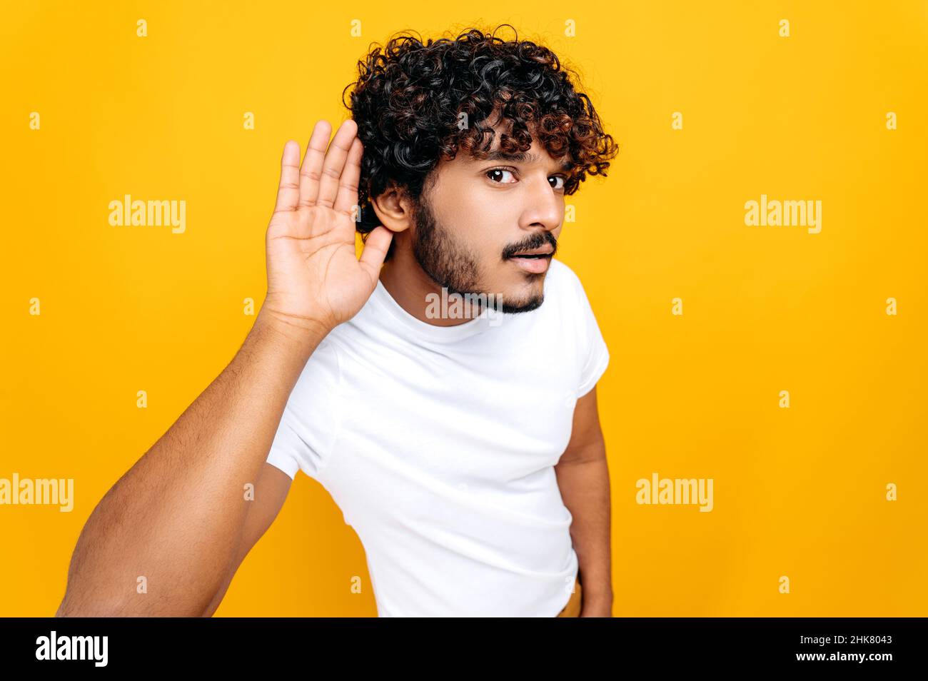 Surdité, potins concept.Homme indien positif, portant un t-shirt de base avec la main près de l'oreille, rumeur d'écoute concentrée, bavardage d'audition, debout sur un fond orange isolé Banque D'Images