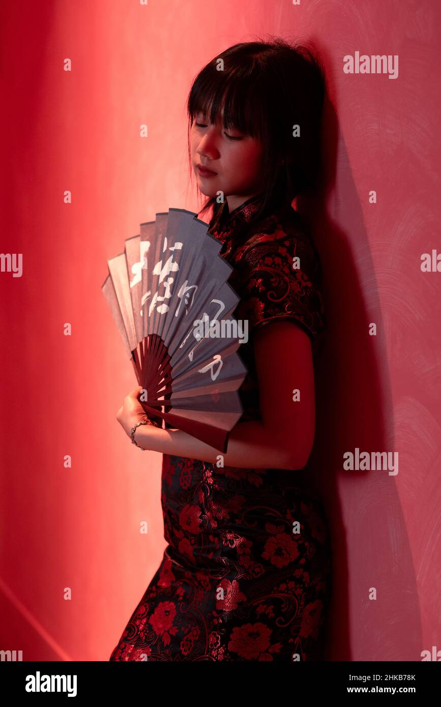 Une fille asiatique est debout contre un mur rouge dans une robe chinoise qipao tenant un ventilateur traditionnel dans ses mains Banque D'Images