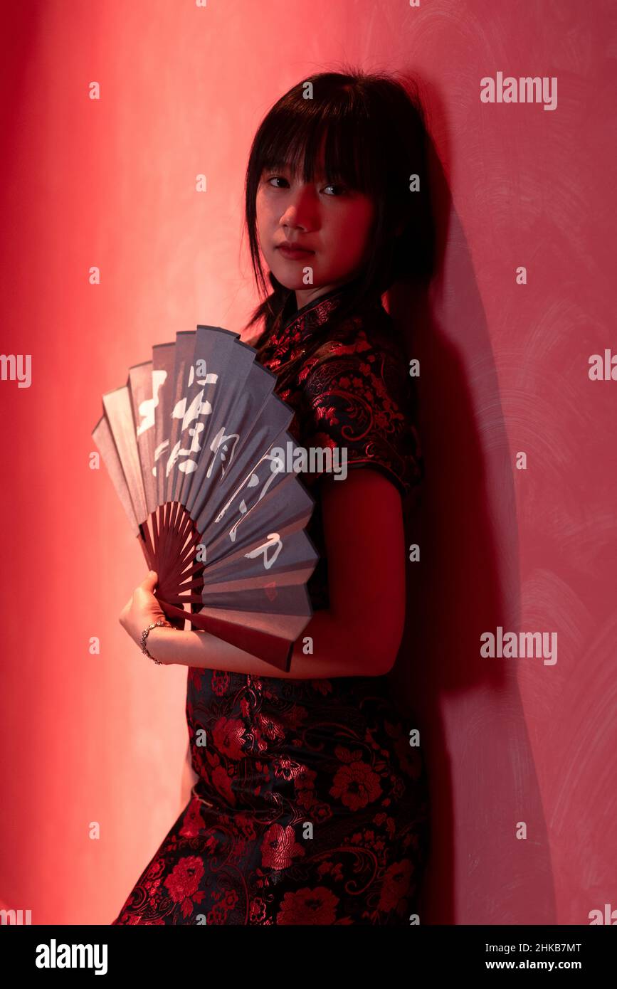 Une fille asiatique est debout contre un mur rouge dans une robe chinoise qipao tenant un ventilateur traditionnel dans ses mains Banque D'Images
