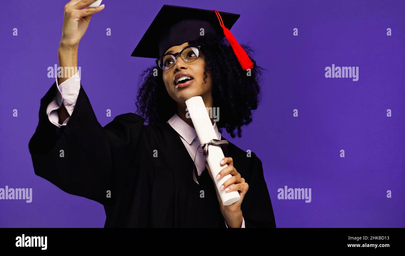 heureux étudiant afro-américain avec des bretelles dans le chapeau de la  remise des diplômes et la robe prenant selfie avec roulé diplôme isolé sur  violet Photo Stock - Alamy