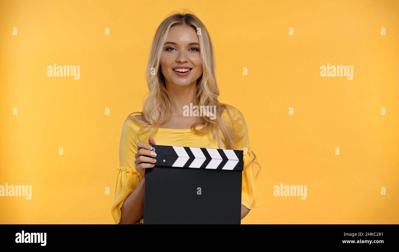 Bonne femme blonde tenant un panneau de clopboard et regardant la caméra isolée sur jaune Banque D'Images