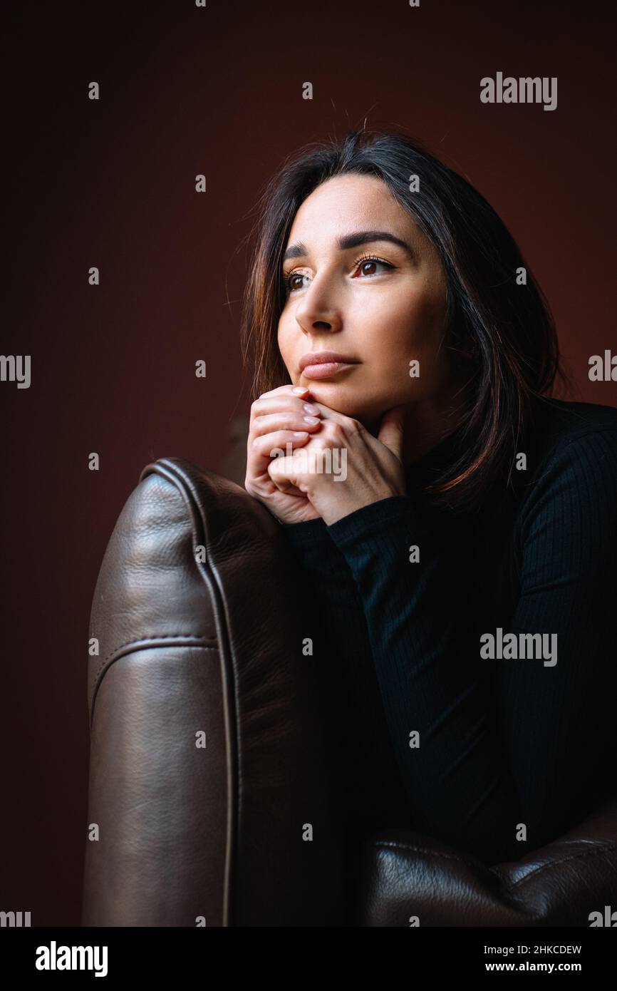 belle femme pensive avec de longs cheveux foncés dans le cou noir de rouleau s'asseoir sur le canapé brun mettant la tête sur les mains claspées. Banque D'Images