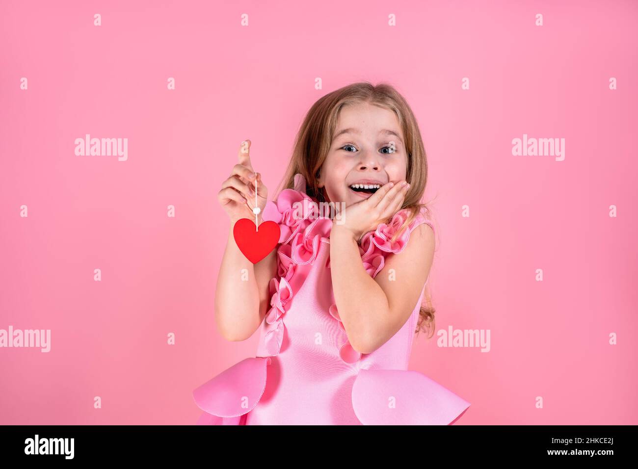 Belle belle petite fille aux yeux bleus avec de longs cheveux en robe rose stand souriant tenant le coeur de papier rouge sur la corde. Banque D'Images