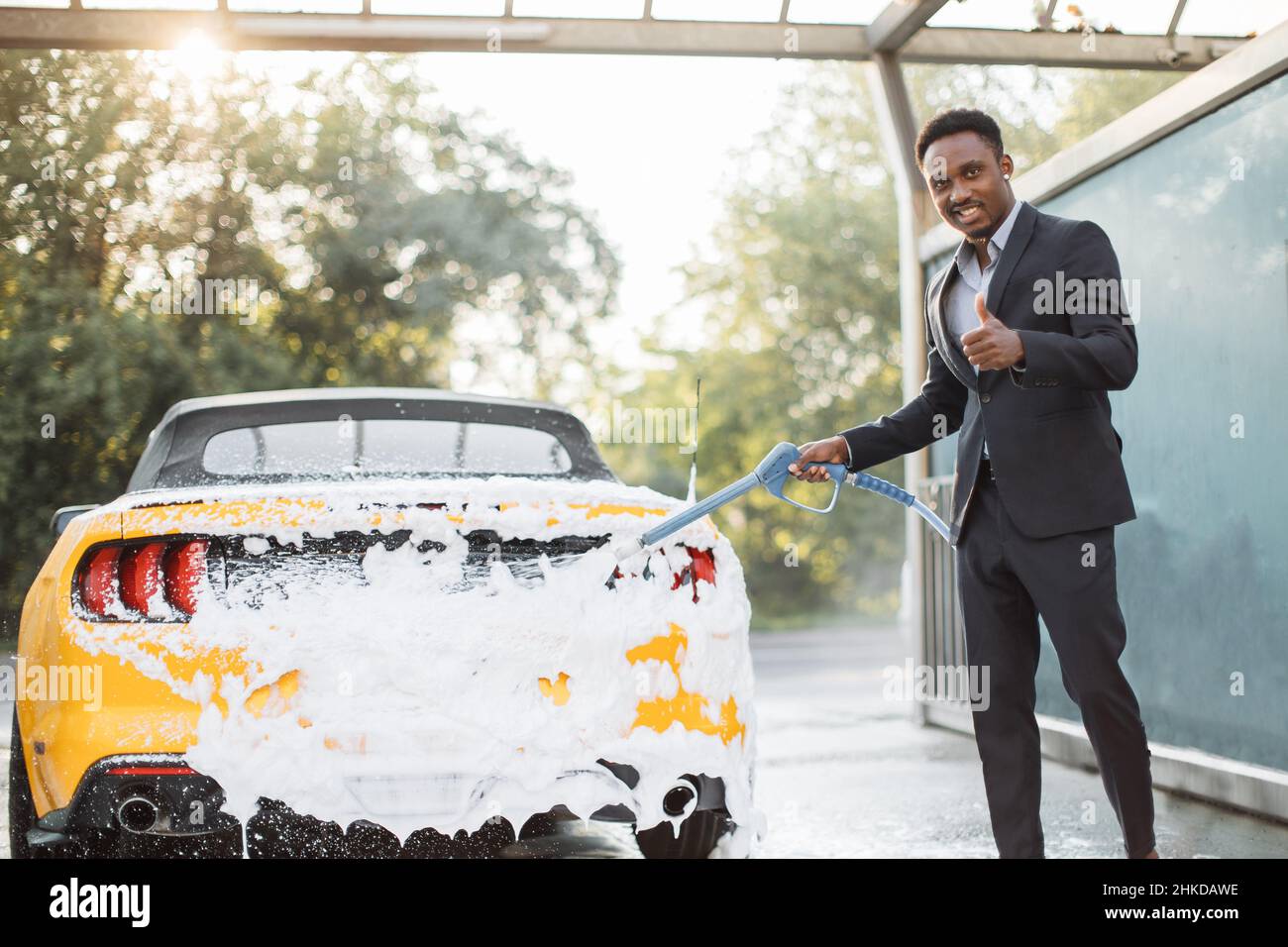 Lavage de voiture moderne jaune sur l'air libre service de lavage de voiture avec mousse et tuyau haute puissance. Jeune homme d'affaires souriant africain confiant nettoyer sa voiture avec un pulvérisateur à jet et du savon à l'extérieur. Banque D'Images