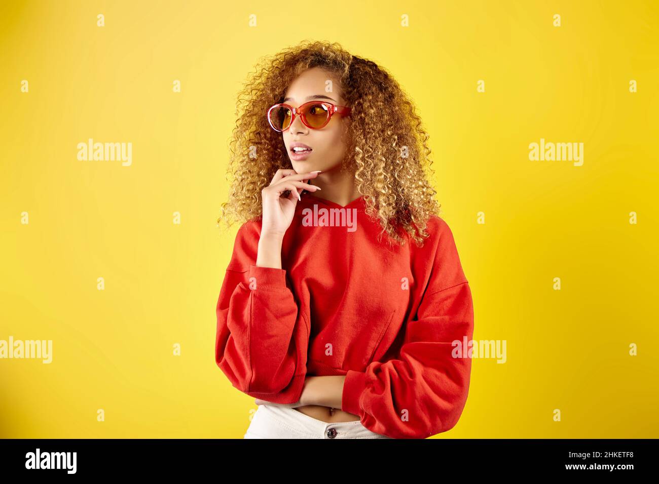 Jeune femme afro-américaine confiante portant un élégant sweat à capuche rouge et des lunettes sur fond jaune en studio Banque D'Images