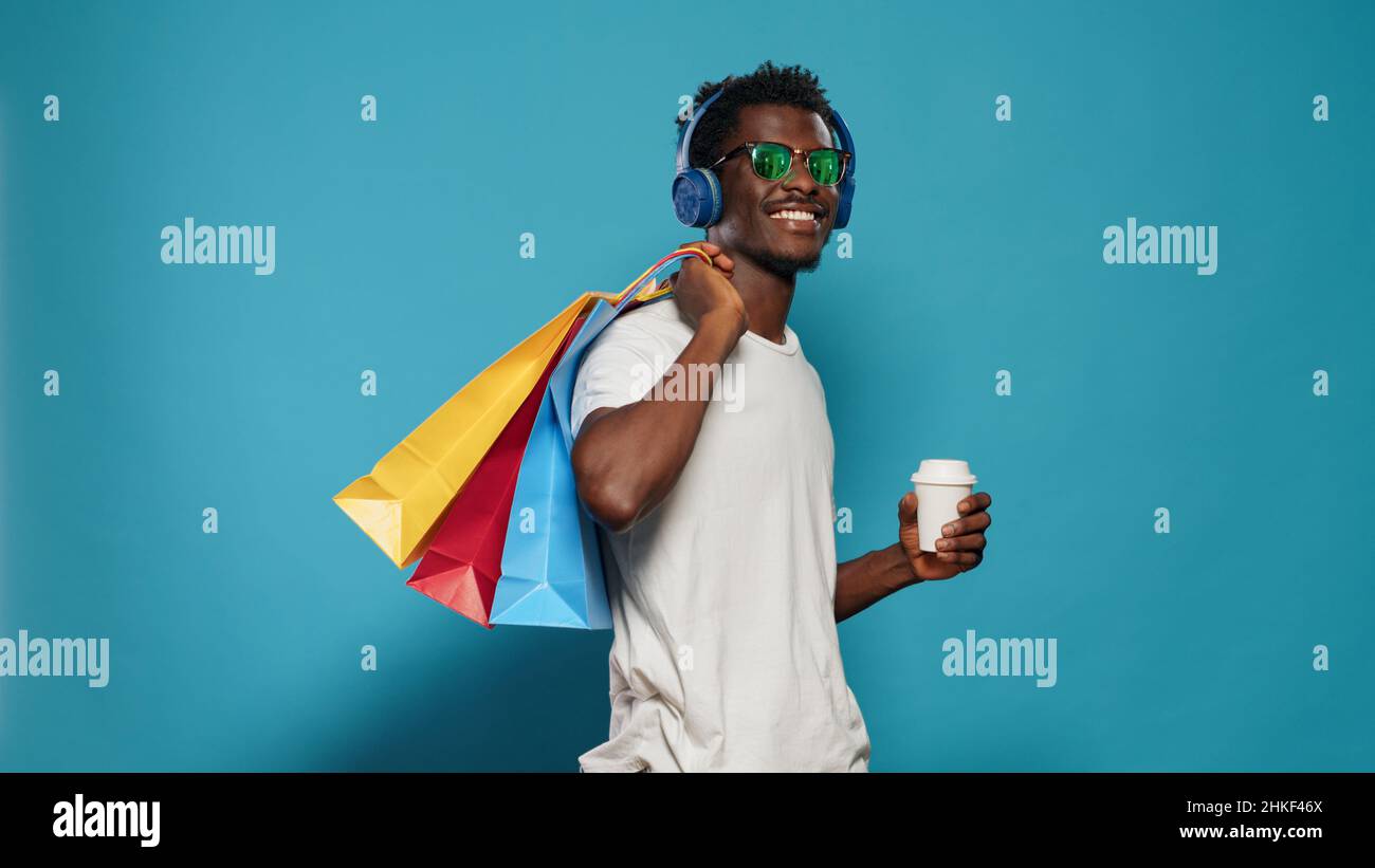 Homme décontracté portant des sacs à provisions et des écouteurs.Personne moderne écoutant de la musique tout en tenant des sacs à main avec des vêtements de magasin et une tasse de café.Shopping adulte Banque D'Images