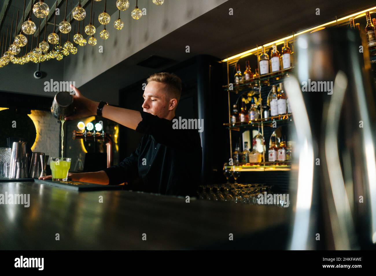 Vue latérale sur le barman qui verse une boisson alcoolisée fraîche dans des verres avec des glaçons derrière le comptoir du bar dans une boîte de nuit moderne. Banque D'Images