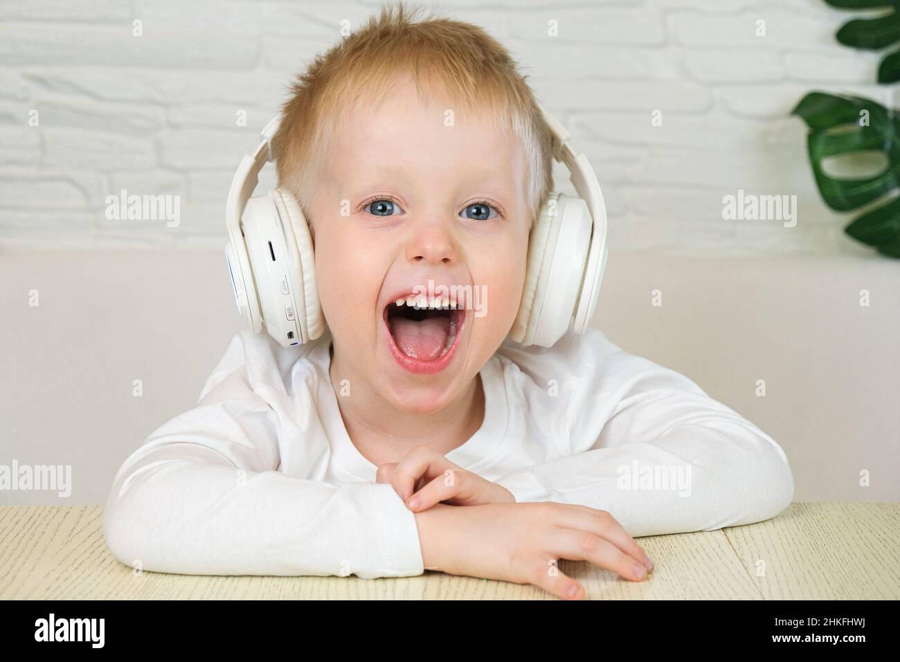 Un petit garçon européen sourit dans un t-shirt blanc avec de grands écouteurs blancs, écoute de la musique et regarde la caméra.Ralenti.Le concept de musique Banque D'Images