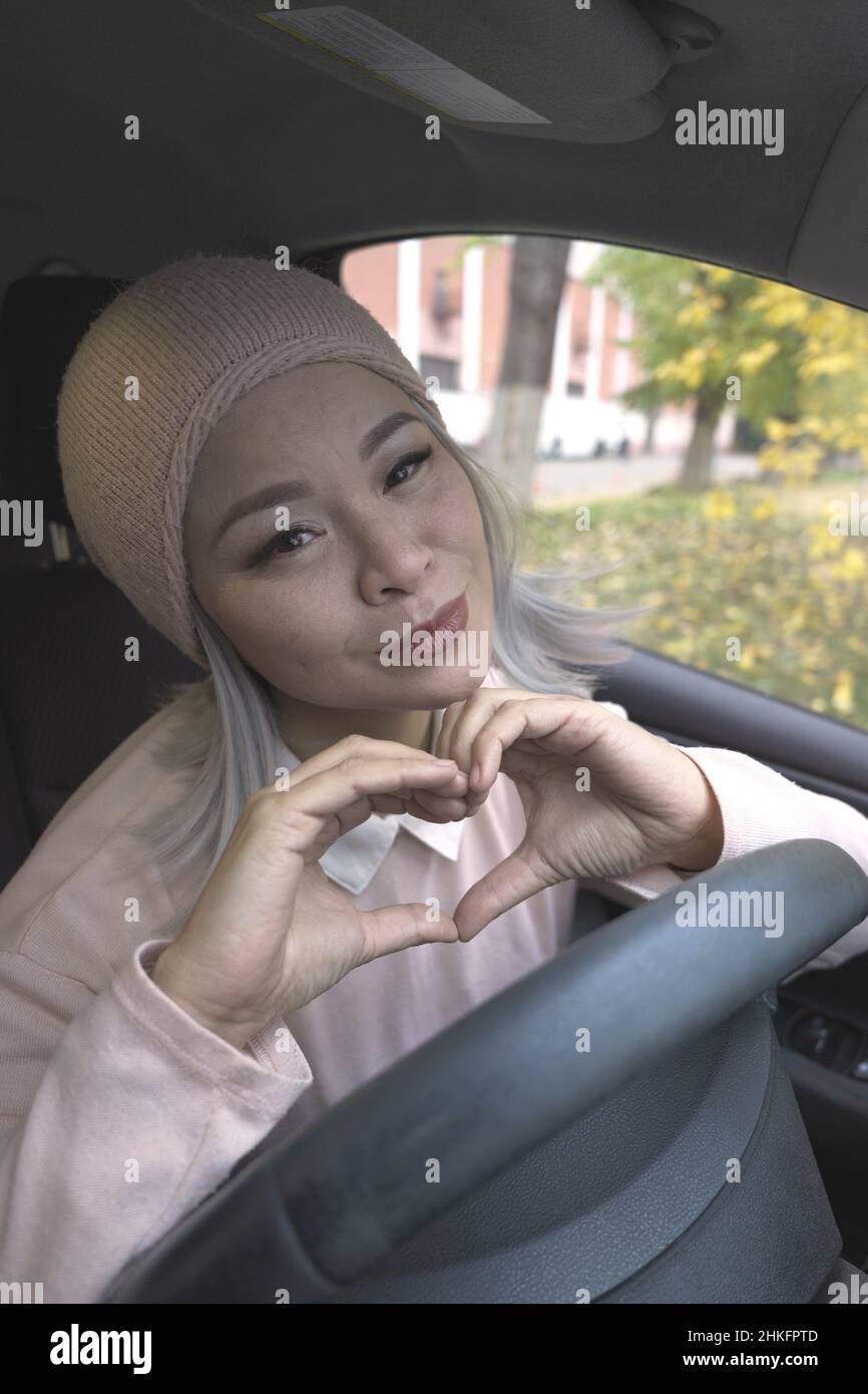 Montrant l'amour blonde femme asiatique dans la voiture.Femme asiatique mature regardant la caméra tenant les mains dans le coeur signe d'amour.Femme asiatique blonde d'âge moyen dans la voiture.Femme sur la route.Concept d'entreprise. Banque D'Images