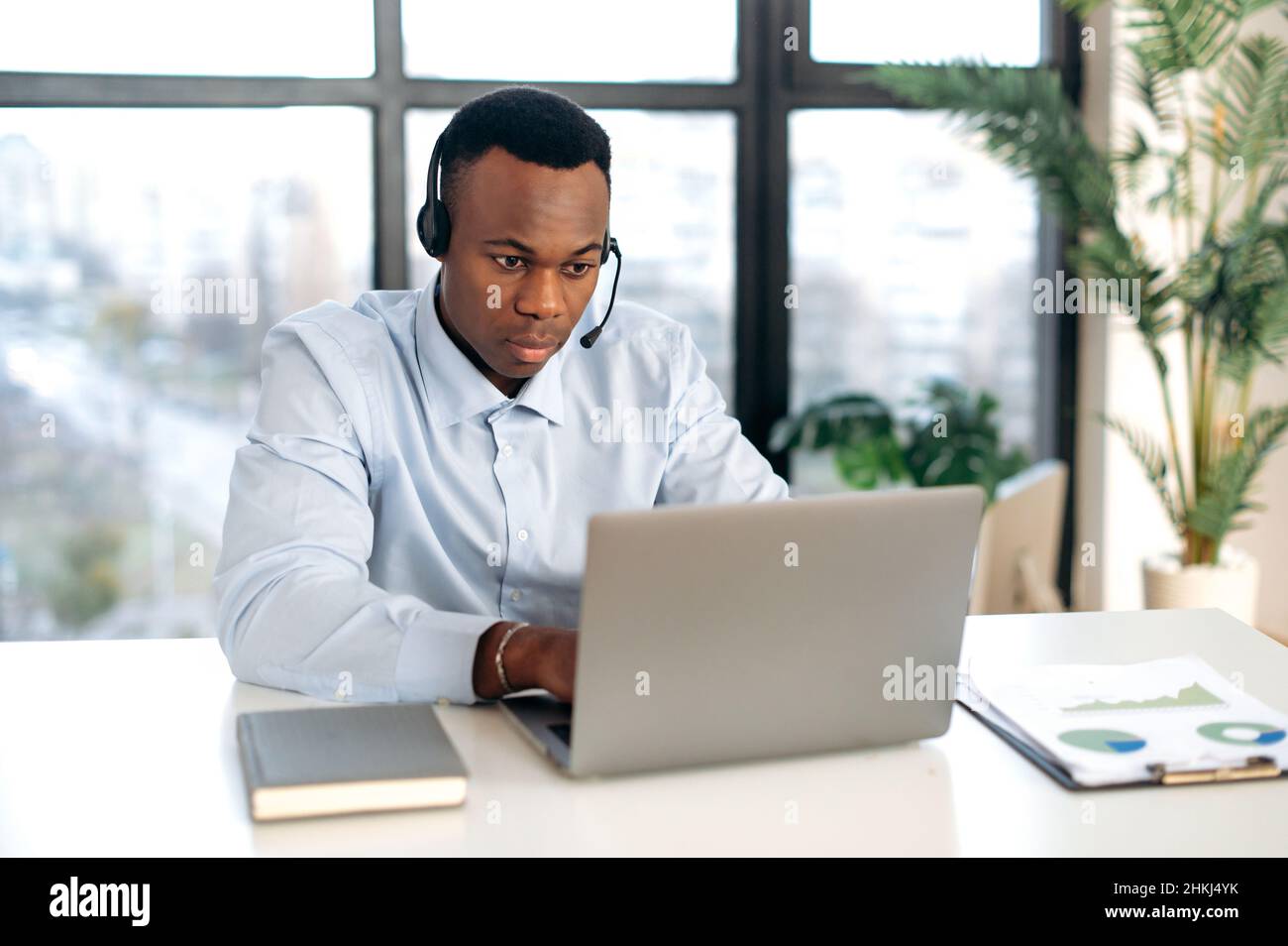 Jeune homme afro-américain intelligent et ciblé dans les casques, le responsable, l'employé d'assistance, le consultant en ligne, assis à un ordinateur portable dans le bureau, ayant une consultation en ligne avec les clients dans une discussion Banque D'Images