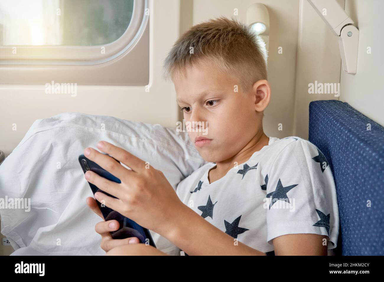 Un adolescent aux cheveux blonds courts lit un livre électronique avec un smartphone allongé sur une literie blanche sur la tablette inférieure du wagon Banque D'Images