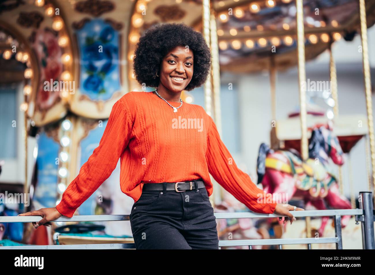 Jeune femme noire souriante avec une coiffure afro et un chandail rouge à côté d'un carrousel dans la rue Banque D'Images